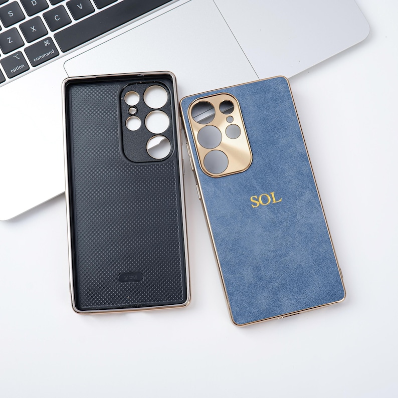 May include: Two phone cases. One is black with a textured pattern and a gold-coloured frame. The other is blue with a gold-coloured frame and the word "SOL" in gold. Both cases are on a white surface.
