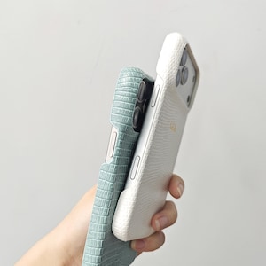 May include: Two smartphone cases, one in a light sage green with a textured crocodile skin pattern and the other in a crisp white with a fine lizard skin texture. Both cases feature precise cutouts for the camera lenses and side buttons.