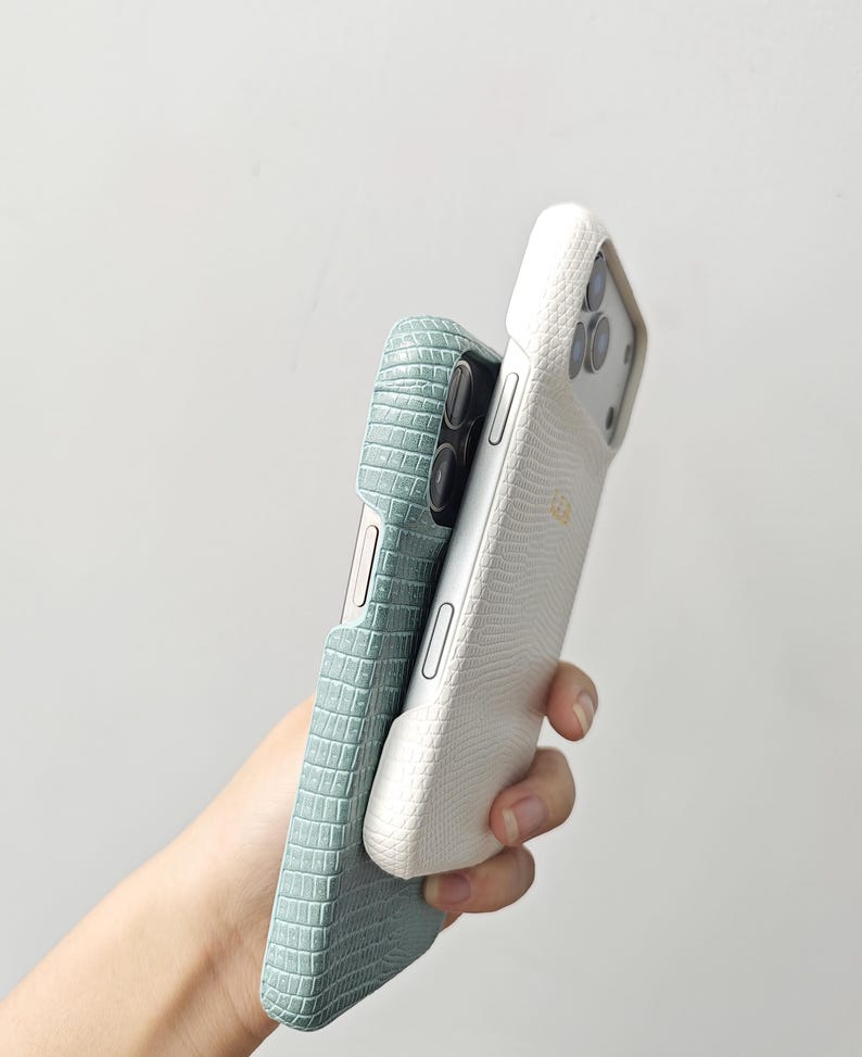 May include: Two smartphone cases, one in a light sage green with a textured crocodile skin pattern and the other in a crisp white with a fine lizard skin texture. Both cases feature precise cutouts for the camera lenses and side buttons.