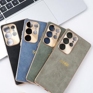 May include: Four smartphone cases in black, blue, green, and grey, each with a gold-coloured camera module and trim. The blue and grey cases have gold lettering. The cases are arranged on a white surface near a laptop.