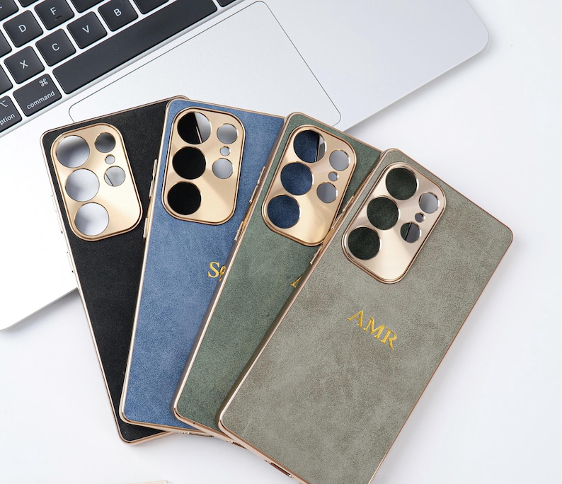 May include: Four smartphone cases in black, blue, green, and grey, each with a gold-coloured camera module and trim. The blue and grey cases have gold lettering. The cases are arranged on a white surface near a laptop.