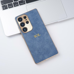 May include: A blue phone case with gold trim and camera housing, featuring the gold letters "SOL". The case is resting on a laptop keyboard.