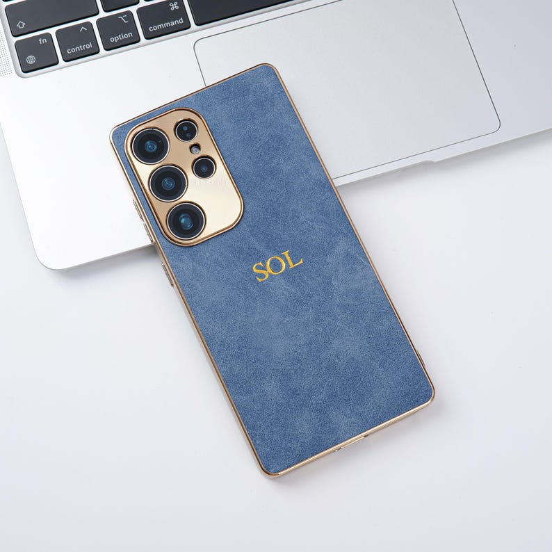 May include: A blue phone case with gold trim and camera housing, featuring the gold letters "SOL". The case is resting on a laptop keyboard.
