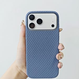 May include: A blue smartphone case with a textured, woven fabric finish. The case features a rectangular cutout at the top for the camera module, which contains three circular lenses and a flash. The material has a distinct diagonal pattern.
