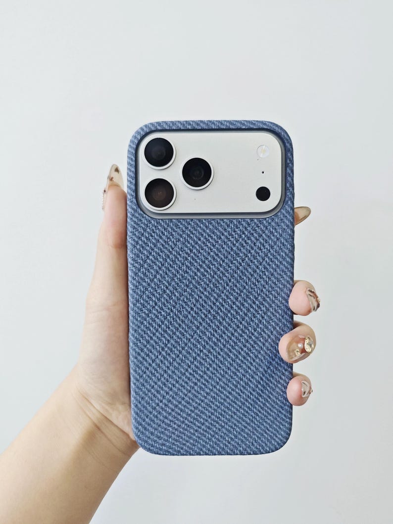 May include: A blue smartphone case with a textured, woven fabric finish. The case features a rectangular cutout at the top for the camera module, which contains three circular lenses and a flash. The material has a distinct diagonal pattern.