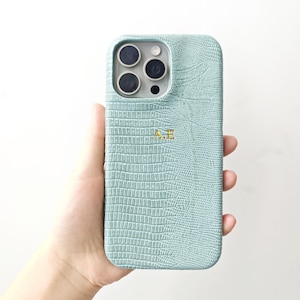 May include: A light mint green phone case with a textured crocodile skin pattern and the gold embossed initials A.E. centred on the back. The case features a precise cutout for the triple camera lens array and flash on the upper left side.
