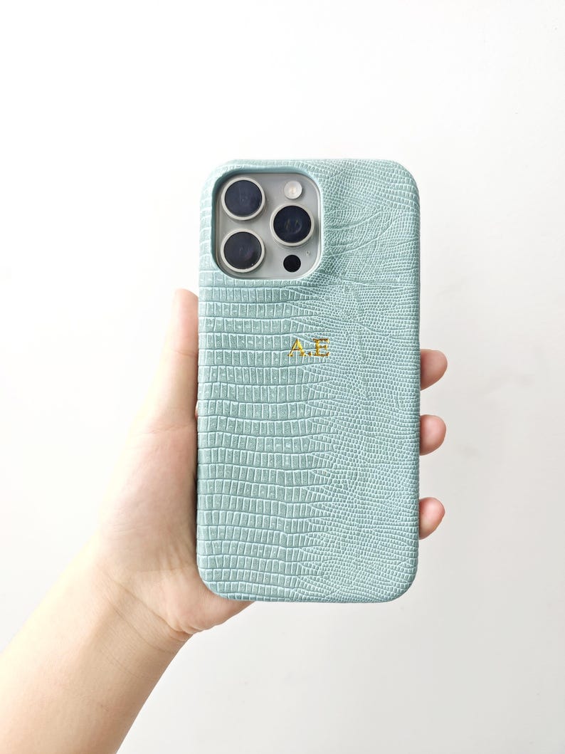 May include: A light mint green phone case with a textured crocodile skin pattern and the gold embossed initials A.E. centred on the back. The case features a precise cutout for the triple camera lens array and flash on the upper left side.