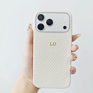 May include: A cream-colored smartphone case with a textured, diagonal woven fabric pattern. The case features the gold-colored initials LD embossed in the center and includes a cutout section at the top for the camera lenses and flash.
