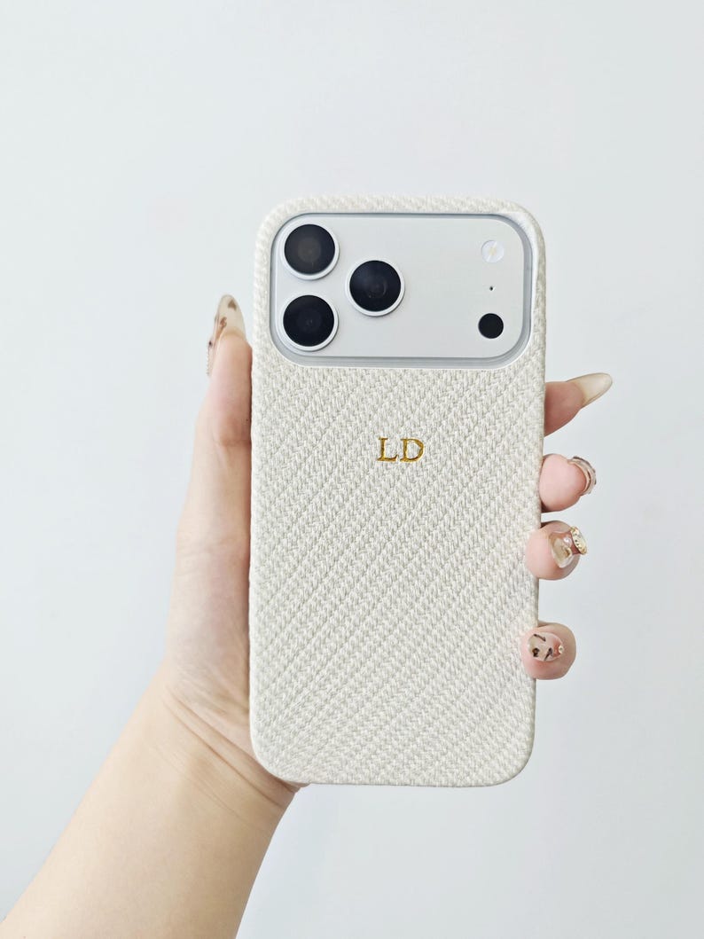 May include: A cream-colored smartphone case with a textured, diagonal woven fabric pattern. The case features the gold-colored initials LD embossed in the center and includes a cutout section at the top for the camera lenses and flash.