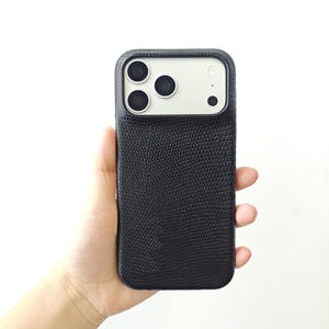 May include: A black smartphone case with a textured, lizard-skin pattern finish. The case features a rectangular cutout at the top to accommodate a triple-lens camera system and a flash, with a smooth, light-coloured panel surrounding the lenses.