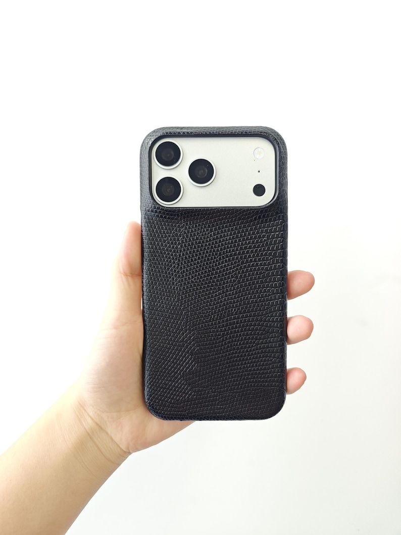 May include: A black smartphone case with a textured, lizard-skin pattern finish. The case features a rectangular cutout at the top to accommodate a triple-lens camera system and a flash, with a smooth, light-coloured panel surrounding the lenses.