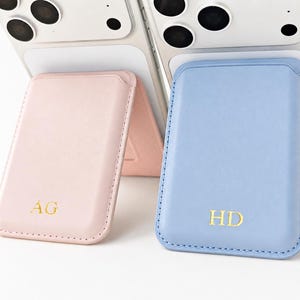 May include: Two rectangular magnetic phone card holders attached to the back of smartphones. One holder is light pink with the gold embossed letters AG, and the other is light blue with the gold embossed letters HD. Both feature stitched edges.