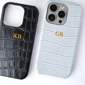 Może przedstawiać: Dwa etui na telefon o fakturze skóry krokodyla. Jedno jest czarne ze złotymi inicjałami "KB", drugie jasnoniebieskie ze złotymi inicjałami "GB". Etui leżą na białej powierzchni.