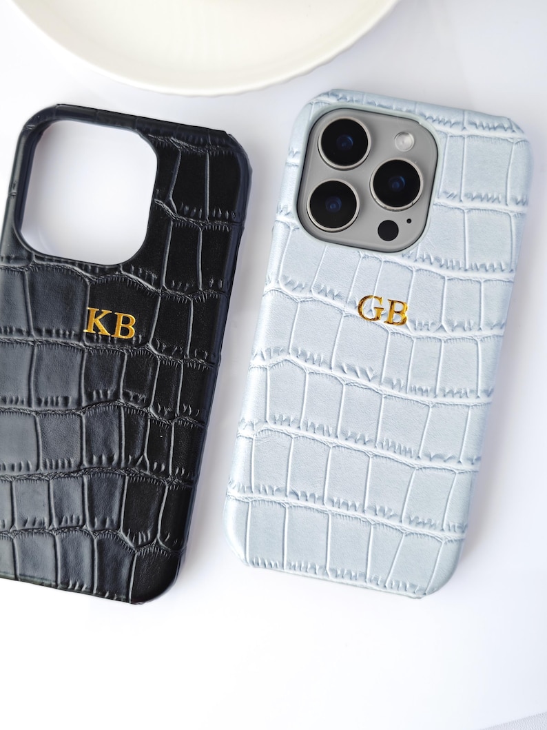 Peut inclure: Deux coques de t&eacute;l&eacute;phone avec une texture peau de crocodile. L'une est noire avec les initiales dor&eacute;es "KB", l'autre est bleu clair avec les initiales dor&eacute;es "GB". Les coques sont sur une surface blanche.