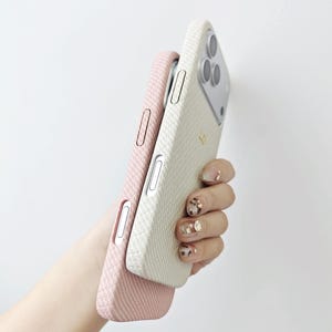 May include: Two smartphone cases with a textured, woven fabric finish. One case is a soft pastel pink, and the other is an off-white cream color. Both cases feature precise cutouts for side buttons and the rear camera module of the mobile device.