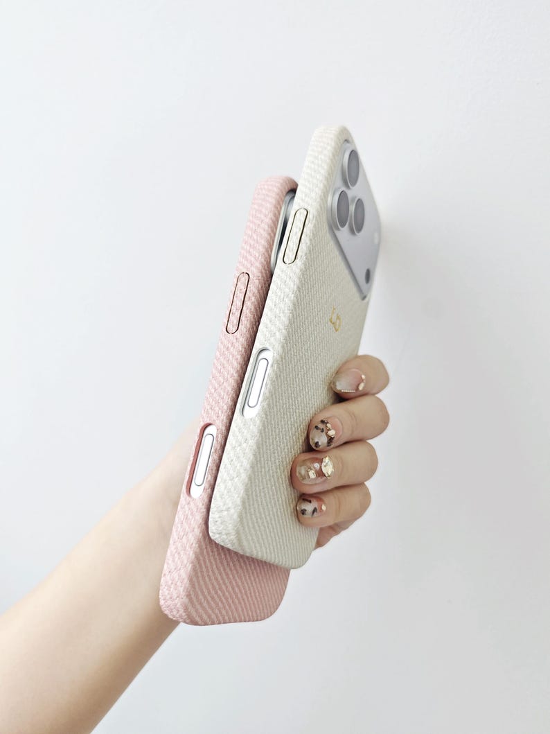 May include: Two smartphone cases with a textured, woven fabric finish. One case is a soft pastel pink, and the other is an off-white cream color. Both cases feature precise cutouts for side buttons and the rear camera module of the mobile device.