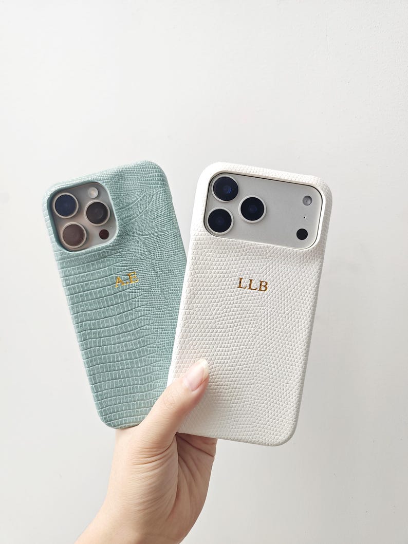 May include: Two smartphone cases with a textured lizard-skin pattern. One case is light sage green with the gold-embossed initials AE, and the other is white with the gold-embossed initials LLB. Both feature cutouts for the camera lenses.