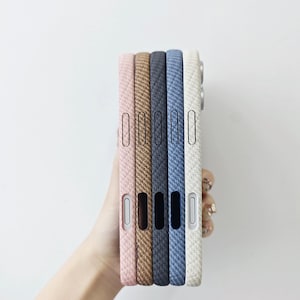 May include: A stack of five smartphone cases featuring a textured, woven fabric-like finish in shades of light pink, tan, dark gray, blue, and off-white. Each case has precise cutouts for side buttons and ports along the vertical edge.