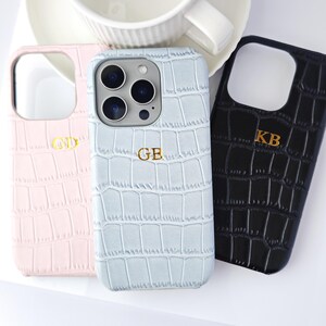 Peut inclure: Trois coques de t&eacute;l&eacute;phone personnalis&eacute;es en rose, bleu clair et noir, chacune avec une texture peau de crocodile. Les coques ont des initiales dor&eacute;es et sont &agrave; c&ocirc;t&eacute; d'une tasse et d'une soucoupe blanches.
