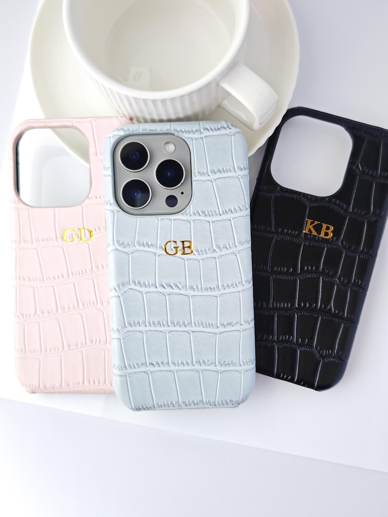 Peut inclure: Trois coques de t&eacute;l&eacute;phone personnalis&eacute;es en rose, bleu clair et noir, chacune avec une texture peau de crocodile. Les coques ont des initiales dor&eacute;es et sont &agrave; c&ocirc;t&eacute; d'une tasse et d'une soucoupe blanches.