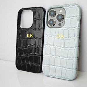 Peut inclure: Deux coques de t&eacute;l&eacute;phone avec une texture peau de crocodile. L'une est noire avec les initiales dor&eacute;es "KB", l'autre est bleu clair avec les initiales dor&eacute;es "GB". Les coques sont con&ccedil;ues pour prot&eacute;ger le t&eacute;l&eacute;phone.