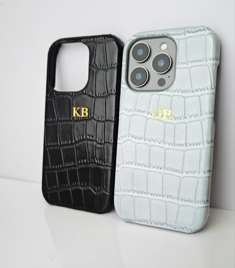Peut inclure: Deux coques de t&eacute;l&eacute;phone avec une texture peau de crocodile. L'une est noire avec les initiales dor&eacute;es "KB", l'autre est bleu clair avec les initiales dor&eacute;es "GB". Les coques sont con&ccedil;ues pour prot&eacute;ger le t&eacute;l&eacute;phone.