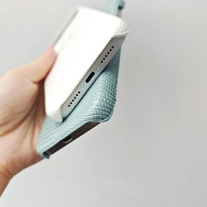 May include: A smartphone case featuring a textured, light blue, lizard-skin pattern on the exterior and a smooth, white, quilted-style finish on the interior edge. The case is shown from an angled perspective, revealing the bottom speaker ports.