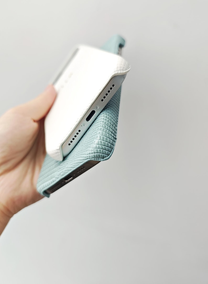 May include: A smartphone case featuring a textured, light blue, lizard-skin pattern on the exterior and a smooth, white, quilted-style finish on the interior edge. The case is shown from an angled perspective, revealing the bottom speaker ports.