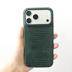 May include: A dark green smartphone case with a textured lizard skin pattern. The case features a rectangular cutout at the top for the camera lenses and flash, revealing a light-coloured phone back underneath the protective cover.