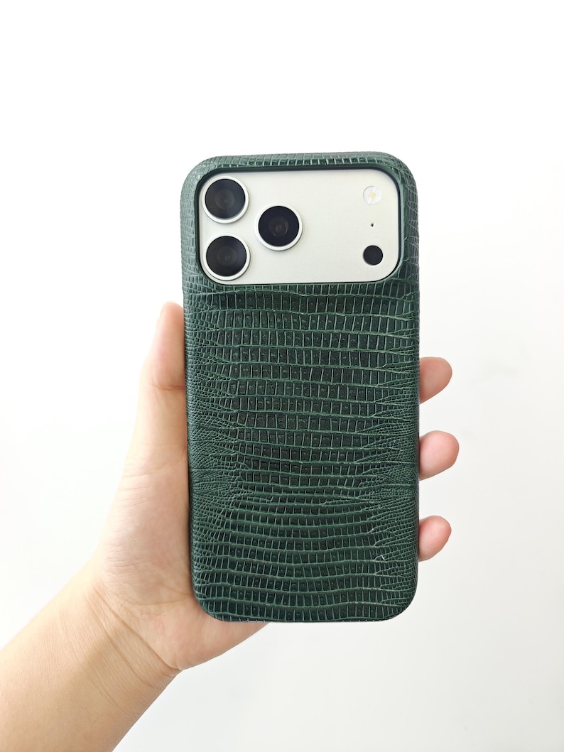 May include: A dark green smartphone case with a textured lizard skin pattern. The case features a rectangular cutout at the top for the camera lenses and flash, revealing a light-coloured phone back underneath the protective cover.