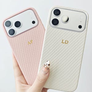 Pode incluir: Duas capas para smartphone com acabamento em tecido texturizado, uma em rosa claro e outra em branco off-white. Cada capa possui um recorte para a câmera e iniciais douradas em relevo, AP na rosa e LD na branca, com um padrão de tecido diagonal.