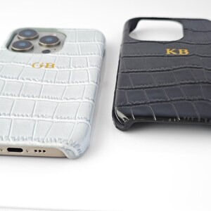 Peut inclure: Deux coques de t&eacute;l&eacute;phone avec une texture peau de crocodile. L'une est gris clair avec les initiales dor&eacute;es "GB", l'autre est noire avec les initiales dor&eacute;es "KB". Les coques sont con&ccedil;ues pour un smartphone.