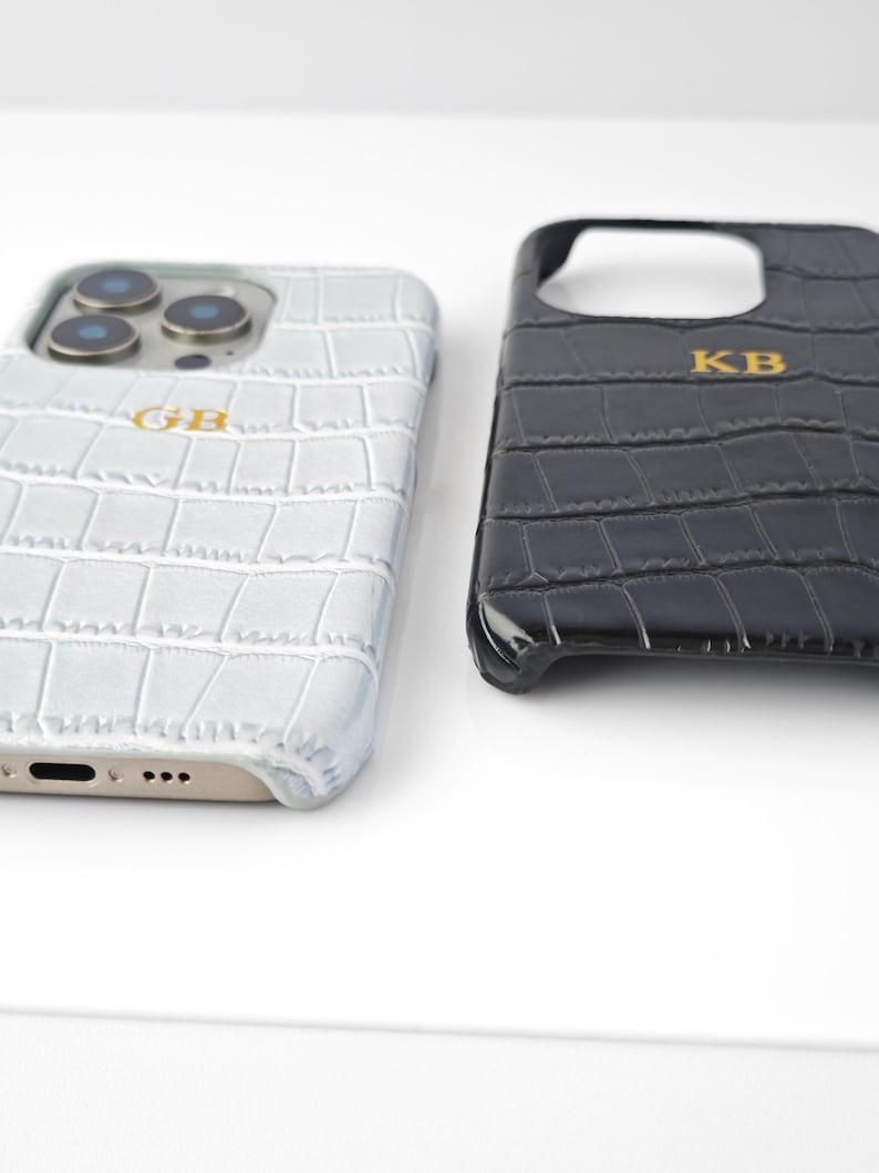 Peut inclure: Deux coques de t&eacute;l&eacute;phone avec une texture peau de crocodile. L'une est gris clair avec les initiales dor&eacute;es "GB", l'autre est noire avec les initiales dor&eacute;es "KB". Les coques sont con&ccedil;ues pour un smartphone.