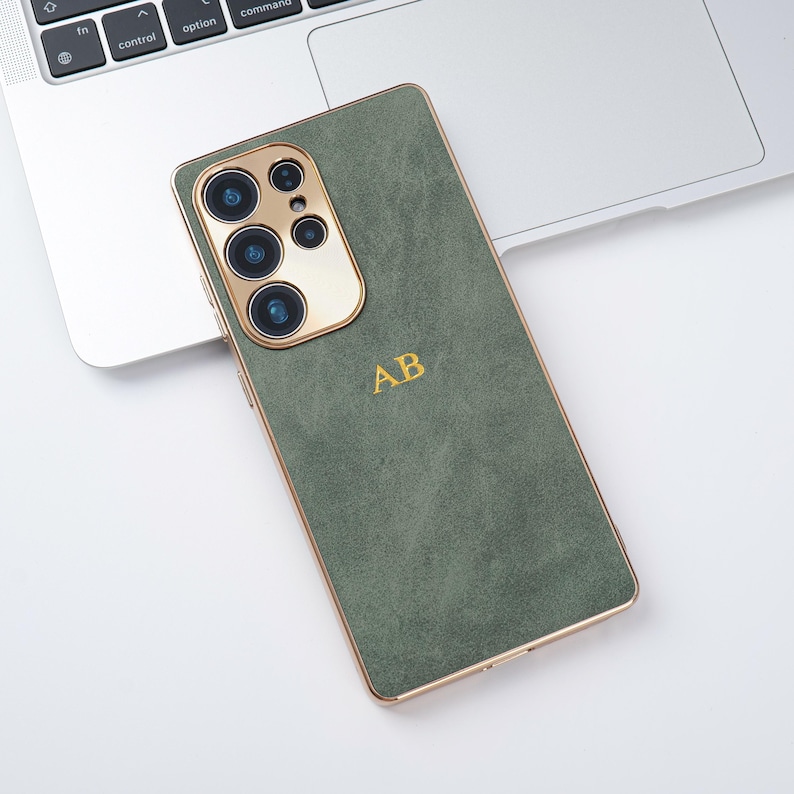 May include: A green phone case with gold edging and a gold camera module. The case features the gold initials "AB". The phone case is on a white surface next to a laptop.