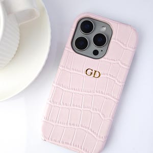 Peut inclure: Coque de t&eacute;l&eacute;phone rose clair avec une texture imitation peau de crocodile. Le bo&icirc;tier porte un monogramme dor&eacute; avec les lettres "GD". Une tasse et une soucoupe blanches sont visibles.