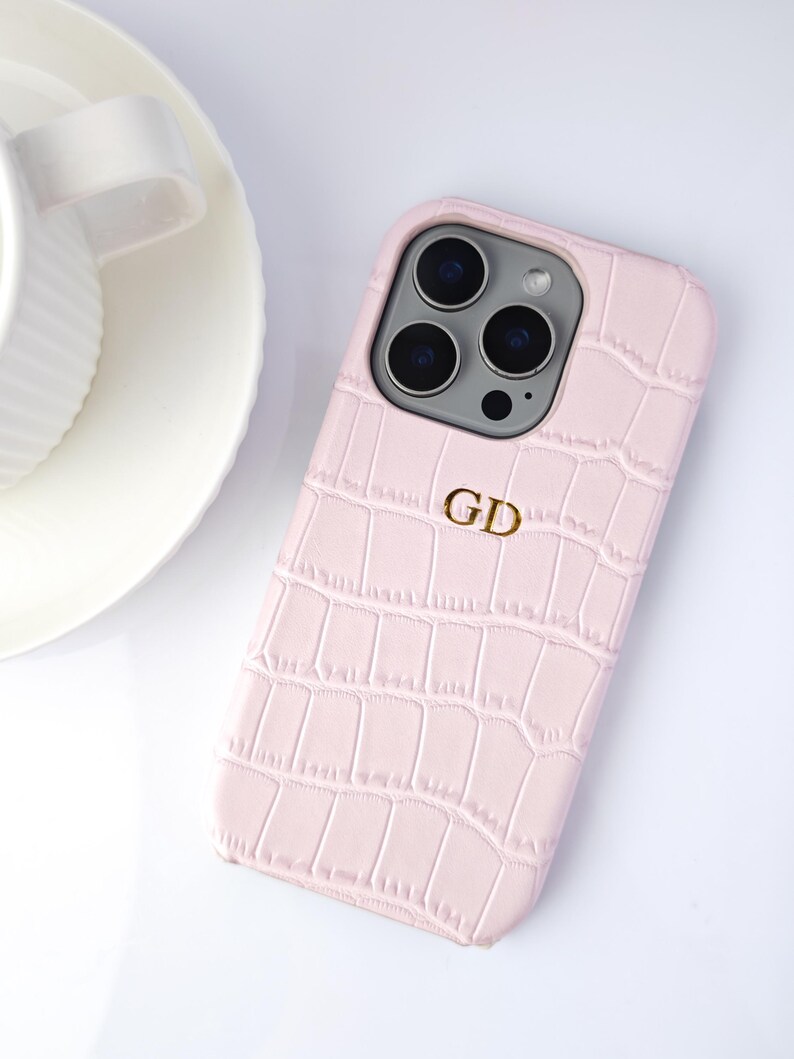 Peut inclure: Coque de t&eacute;l&eacute;phone rose clair avec une texture imitation peau de crocodile. Le bo&icirc;tier porte un monogramme dor&eacute; avec les lettres "GD". Une tasse et une soucoupe blanches sont visibles.