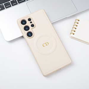 May include: A cream-coloured phone case with a textured finish, featuring a circular design with the gold letters "CD". The case is next to a laptop and a small spiral-bound notebook.