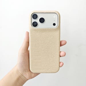 May include: A beige smartphone case with a textured lizard skin pattern. The case features a rectangular cutout at the top for the camera module, which contains three circular lenses and a small circular flash, all set against a light grey background.