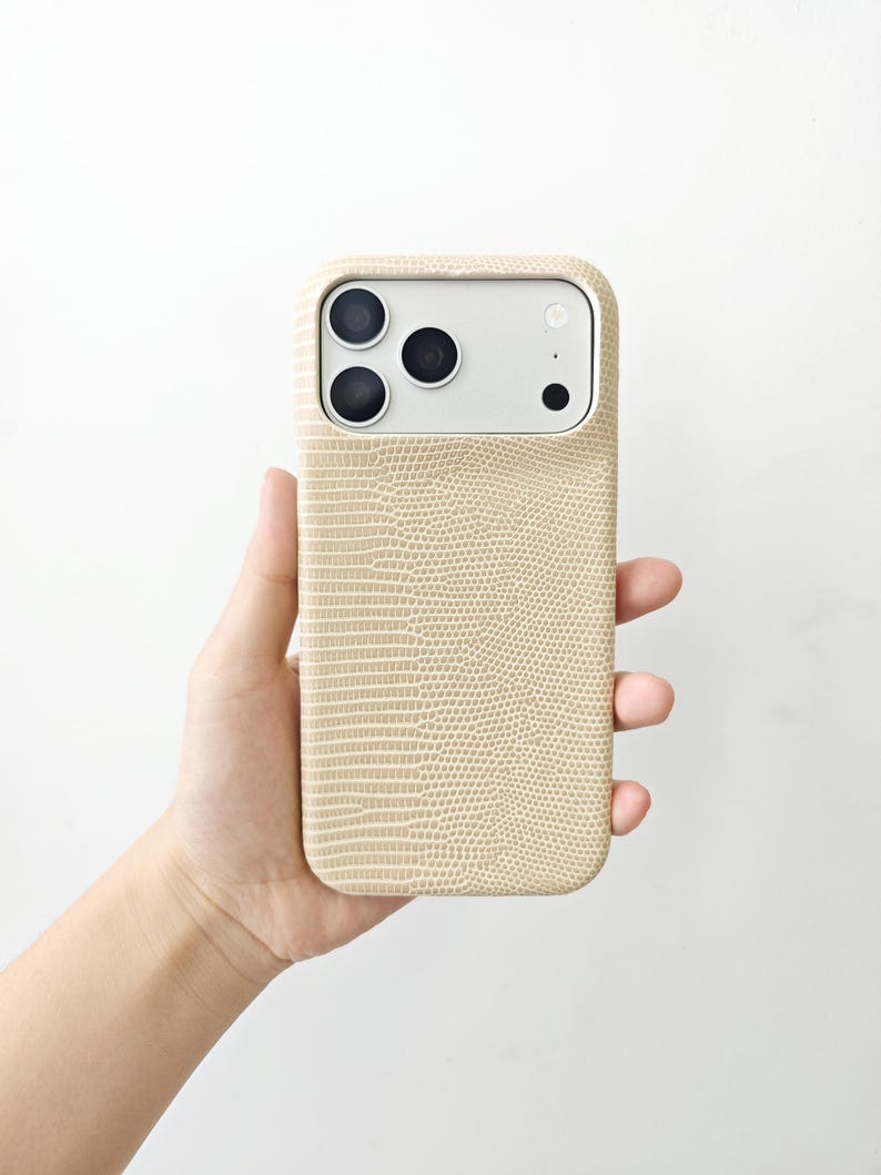 May include: A beige smartphone case with a textured lizard skin pattern. The case features a rectangular cutout at the top for the camera module, which contains three circular lenses and a small circular flash, all set against a light grey background.