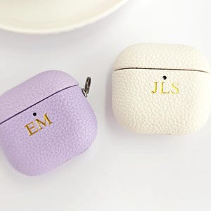 May include: Two textured earbud cases with a metal side loop. One case is light purple with the gold embossed letters EM, and the other is off-white with the gold embossed letters JLS. Both cases feature a small circular cutout for a status light.