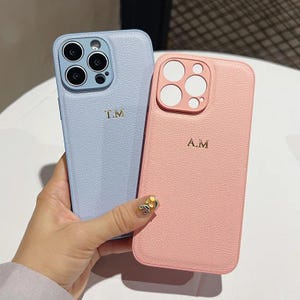 May include: Two personalized phone cases. One is light blue with the initials "T.M." in gold. The other is pink with the initials "A.M." in gold. Both cases have a textured leather-like appearance.