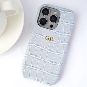 Peut inclure: Coque de t&eacute;l&eacute;phone bleu clair avec une texture imitation peau de crocodile. Le bo&icirc;tier porte un monogramme dor&eacute; avec les lettres "GB". La coque est pos&eacute;e sur une surface blanche.