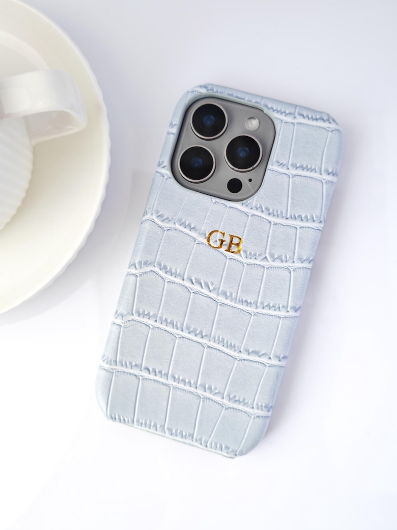 Peut inclure: Coque de t&eacute;l&eacute;phone bleu clair avec une texture imitation peau de crocodile. Le bo&icirc;tier porte un monogramme dor&eacute; avec les lettres "GB". La coque est pos&eacute;e sur une surface blanche.