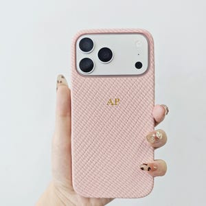 May include: A light pink smartphone case with a textured herringbone fabric finish. The case features a rectangular camera cutout with three circular lenses and a flash. The letters AP are embroidered in gold thread in the center of the case.
