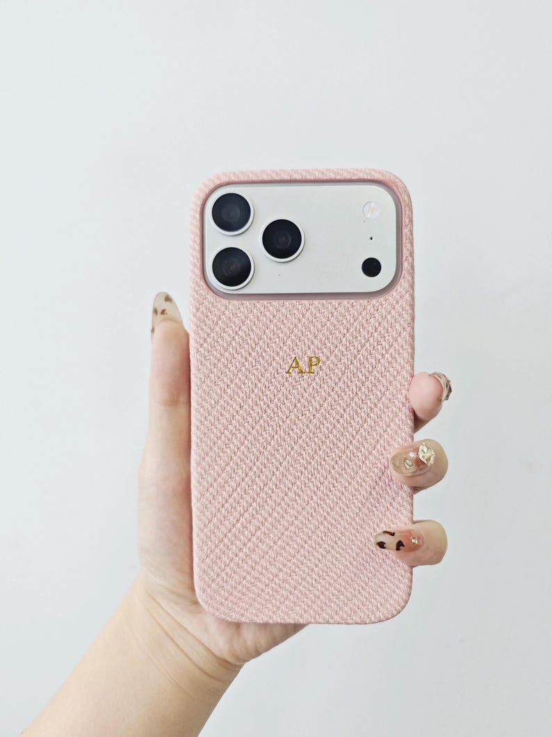 May include: A light pink smartphone case with a textured herringbone fabric finish. The case features a rectangular camera cutout with three circular lenses and a flash. The letters AP are embroidered in gold thread in the center of the case.