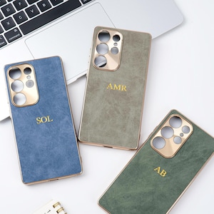 May include: Three personalised phone cases in blue, green, and olive green, each with a gold-coloured camera cutout and trim. The cases are embossed with gold initials: SOL, AMR, and AB. The cases are displayed on a white surface near a laptop.