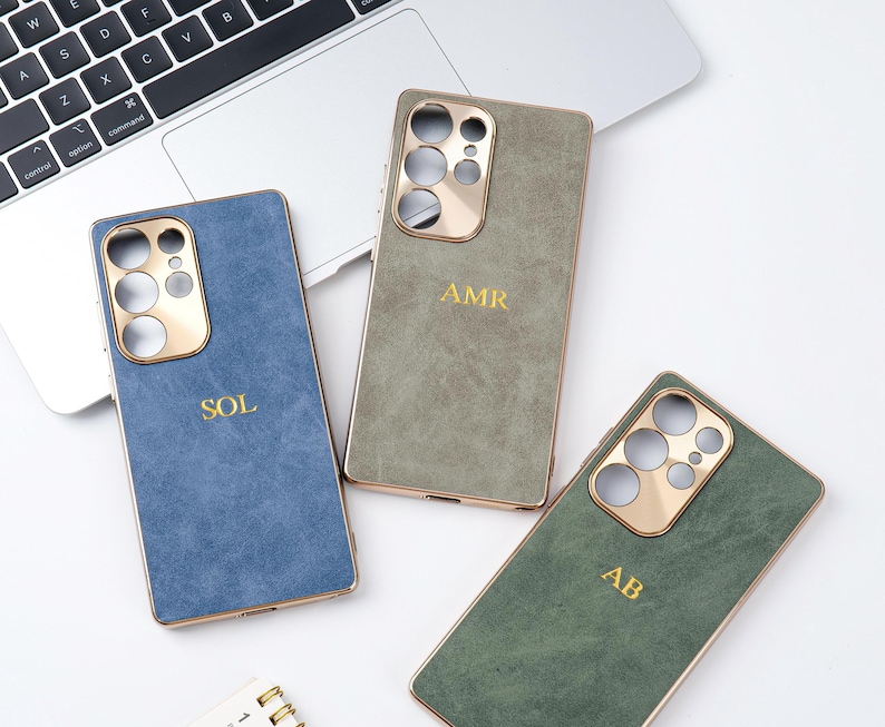 May include: Three personalised phone cases in blue, green, and olive green, each with a gold-coloured camera cutout and trim. The cases are embossed with gold initials: SOL, AMR, and AB. The cases are displayed on a white surface near a laptop.