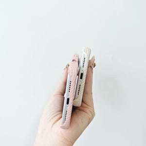 May include: Two mobile phone cases with a textured surface, one in a soft pink shade and the other in a cream white color. Both cases feature precise cutouts for the charging port and speaker grilles along the bottom edge of the devices.
