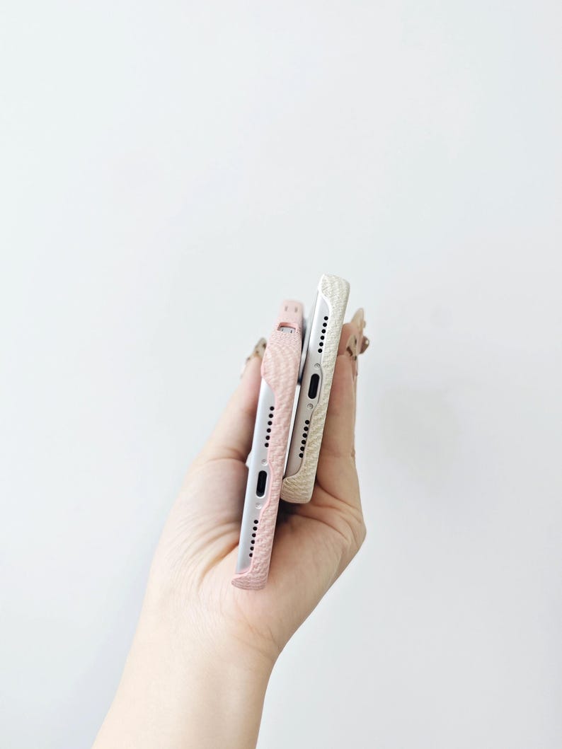 May include: Two mobile phone cases with a textured surface, one in a soft pink shade and the other in a cream white color. Both cases feature precise cutouts for the charging port and speaker grilles along the bottom edge of the devices.