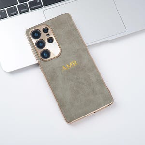 May include: A sage green phone case with gold trim and a gold camera module. The case features the initials "AMR" in gold lettering. The phone is on a white surface next to a laptop.